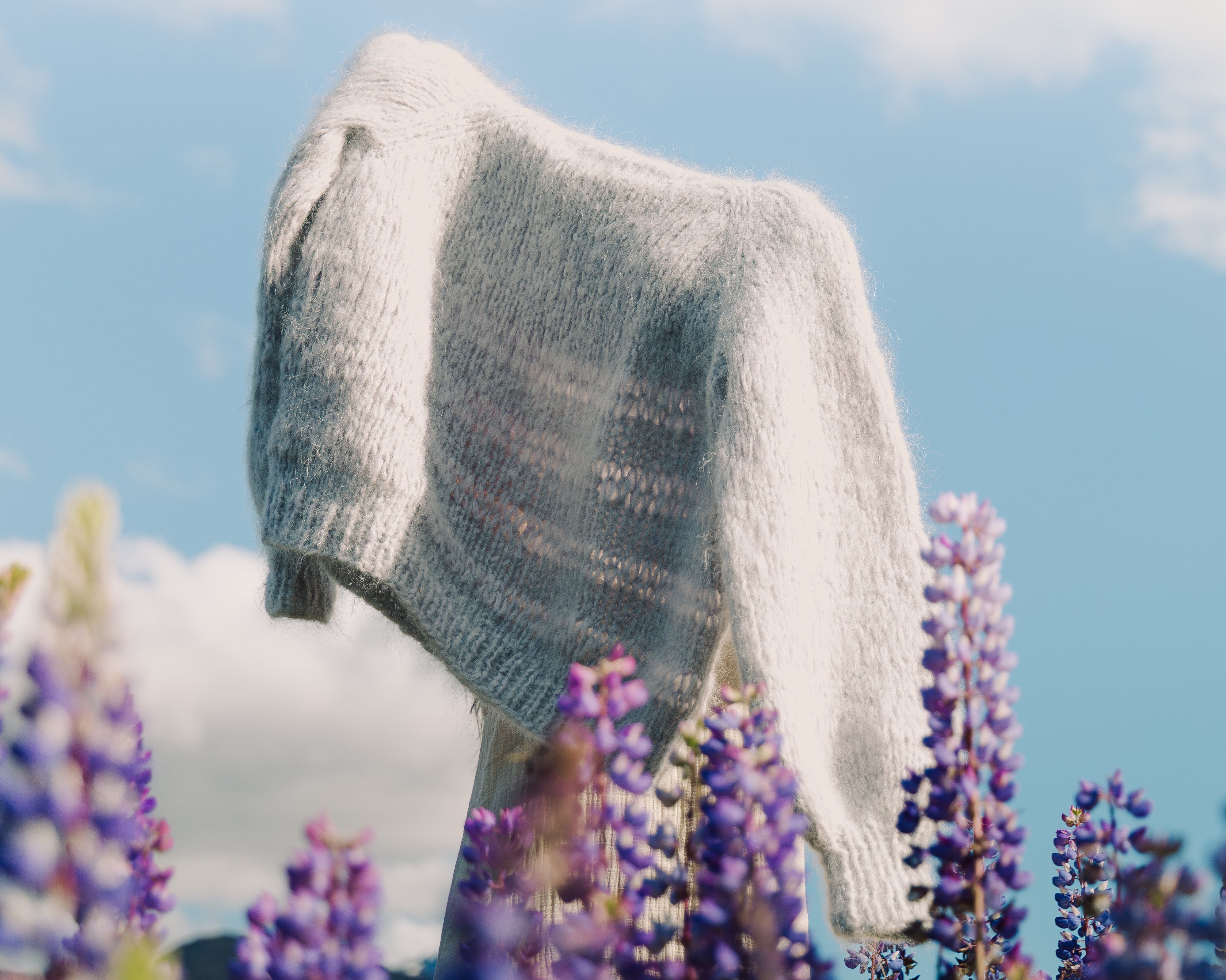 A light gray knit sweater draped outdoors against a blue sky with clouds, surrounded by purple lupin flowers in the foreground.
